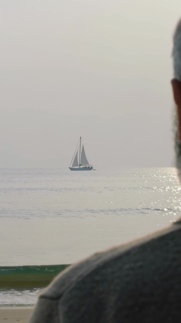 Video verticale: Entra da destra un uomo anziano che indossa un maglione grigio, che osserva una barca a vela che va alla deriva.
