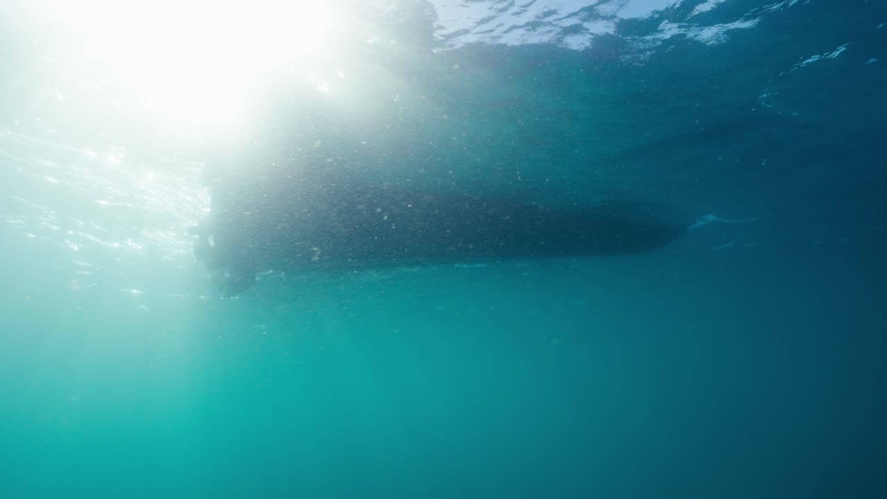 Underwater clip showing the hull of a boat as it floats on the ocean surface with the suns rays shining through the water