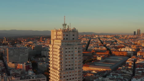 Fly-around-historic-high-rise-building-with-balconies,-houses-in-urban-district-in-background.-Illuminated-by-setting-sun.