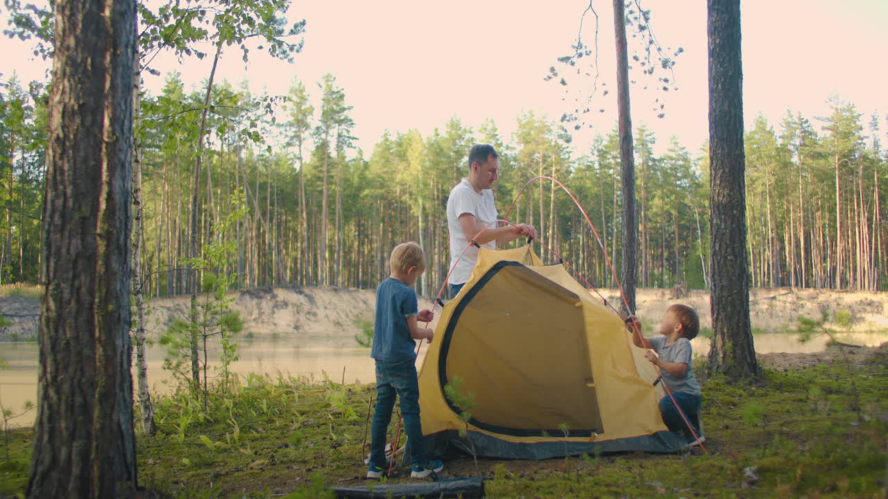 los niños junto con su padre establecen una tienda de campaña para la noche y acampan en el bosque durante el viaje. un hombre y dos niños de 3-5 años juntos en una caminata reúnen una tienda de campaña en cámara lenta