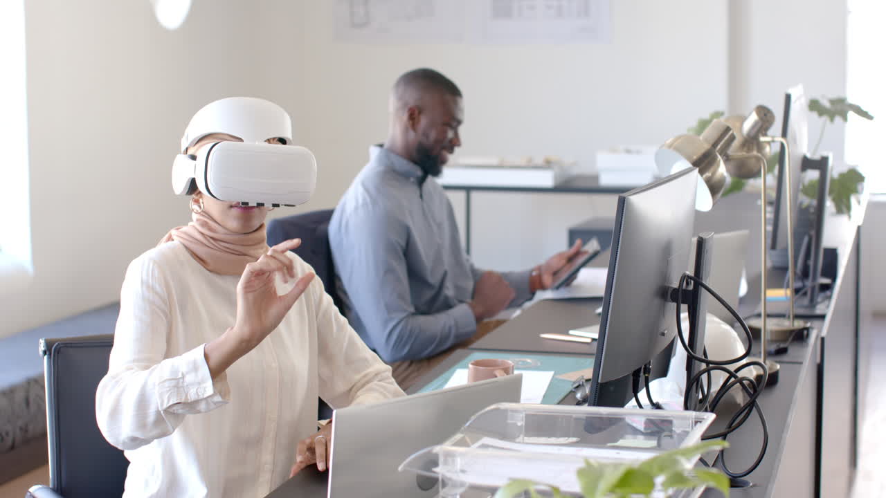 Using VR headset, woman working at desk while colleague uses tablet in office