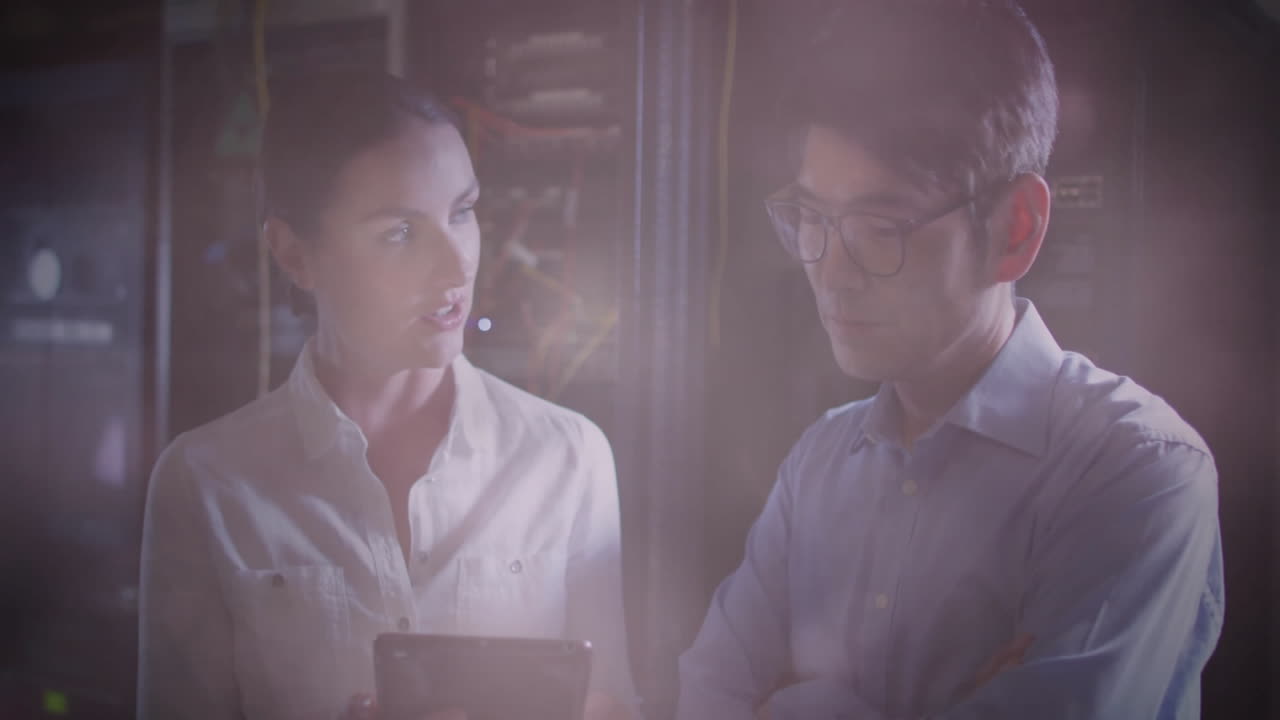 Woman IT professional holding tablet, man studying holographic data with virtual UI in server room
