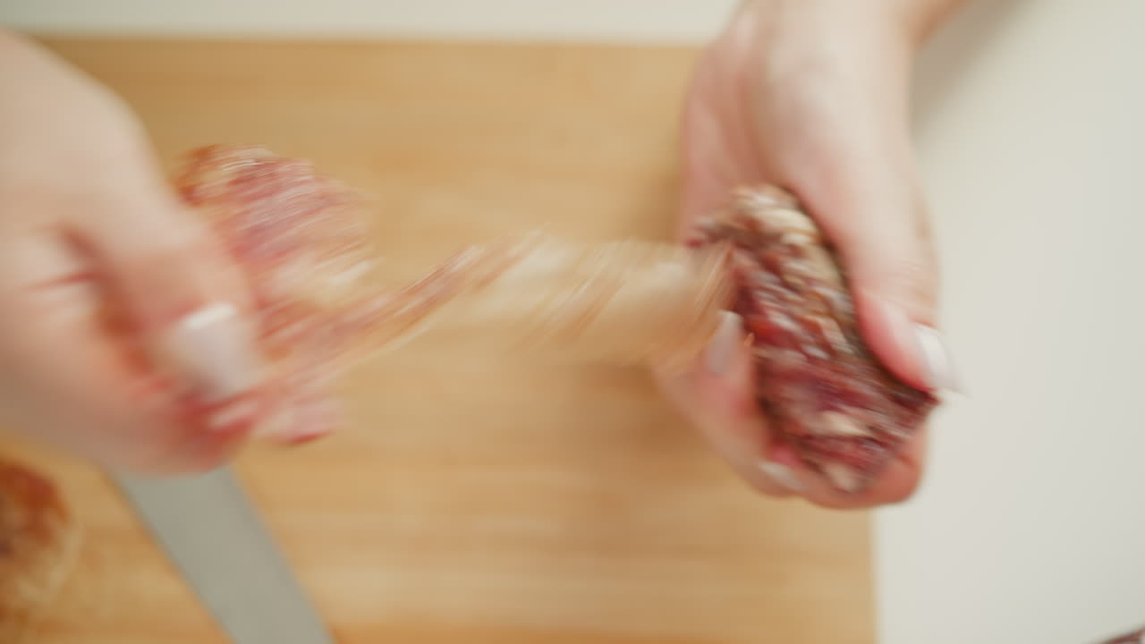 Fair skinned lady using fingers to peel casing from cured sausage over wooden board on white table under soft window light, showcasing tactile food prep and textured meat removal technique