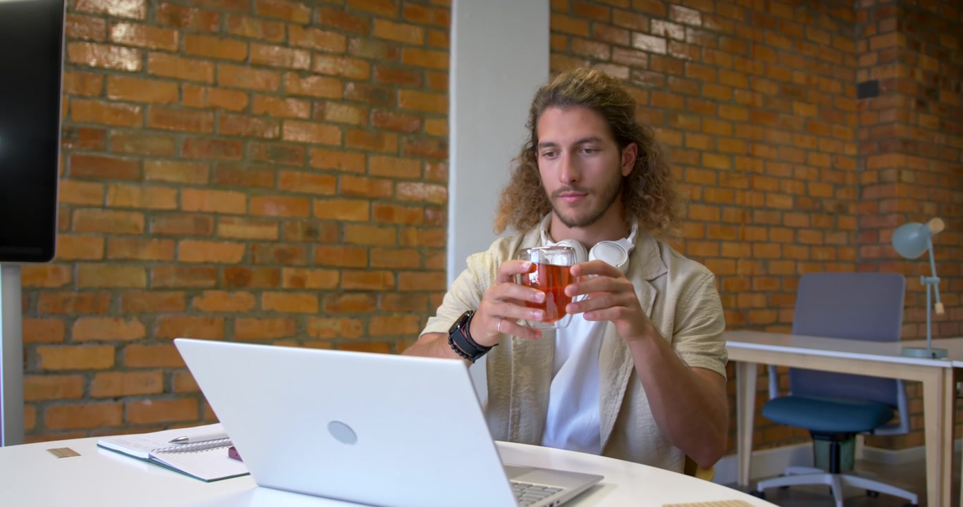 Digita sul laptop, l'uomo guarda la tazza, sistema il sottobicchiere, sorseggia il tè per rinfrescarsi alla scrivania bianca