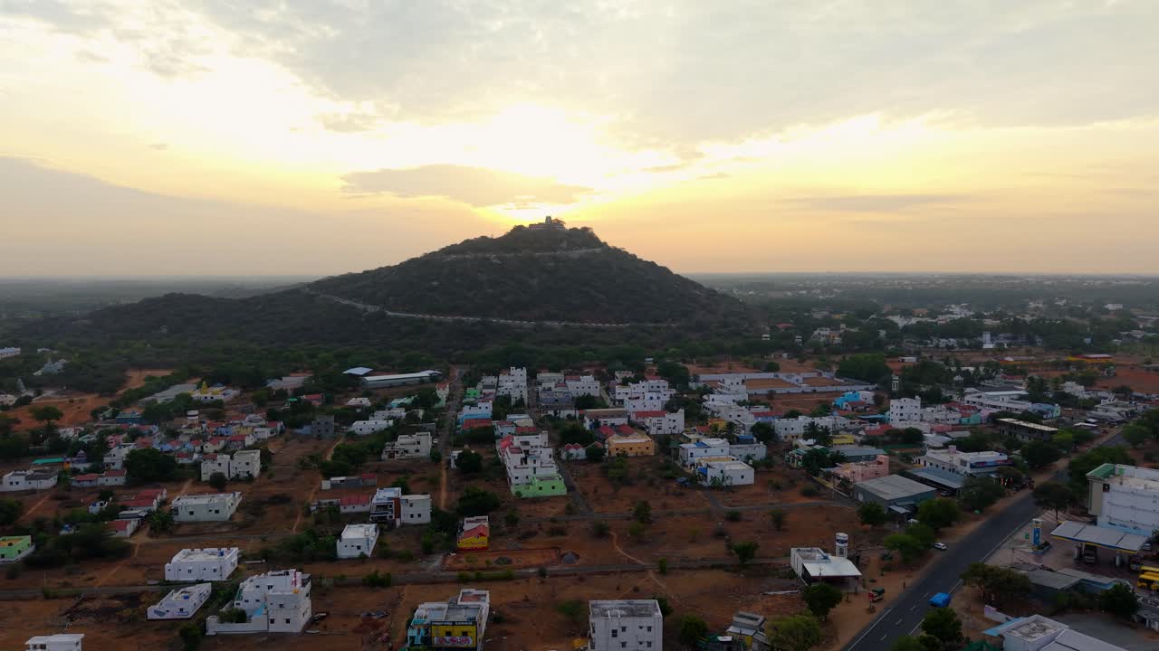 A stunning aerial sunrise view of a hilltop temple with golden rays lighting the sky, surrounded by lush greenery and a small town with colorful houses at the foothills, showcasing natural beauty
