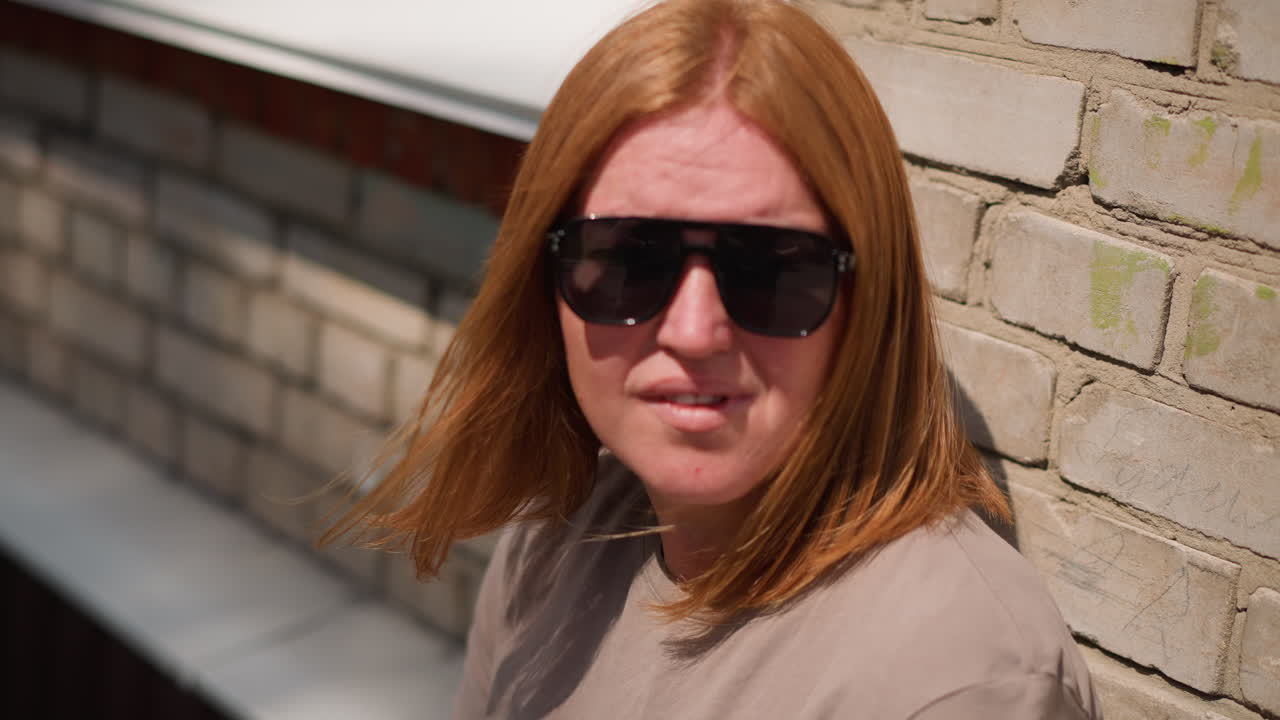 Donna con capelli castano chiaro appoggiata a un muro di mattoni in una giornata di sole, che si sistema gli occhiali da sole mentre guarda a destra e a sinistra, con la calda luce del sole che illumina la pelle e le emozioni