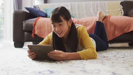 Chica-Asiática-Sonriendo-Y-Usando-Una-Tableta-Tirada-En-El-Suelo-En-Casa