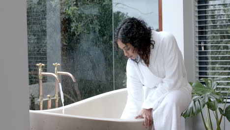 biracial woman wearing bathrobe sitting on bathtub running bath in bathroom at home, slow motion