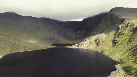 Hermosa-Antena-De-Un-Pequeño-Río-Con-Un-Lago-Dentro-De-Montañas-Cubiertas-De-Hierba-Con-Una-Cordillera-Cubierta-De-Nubes-Pedregosas-Que-Se-Encuentra-Detrás-2