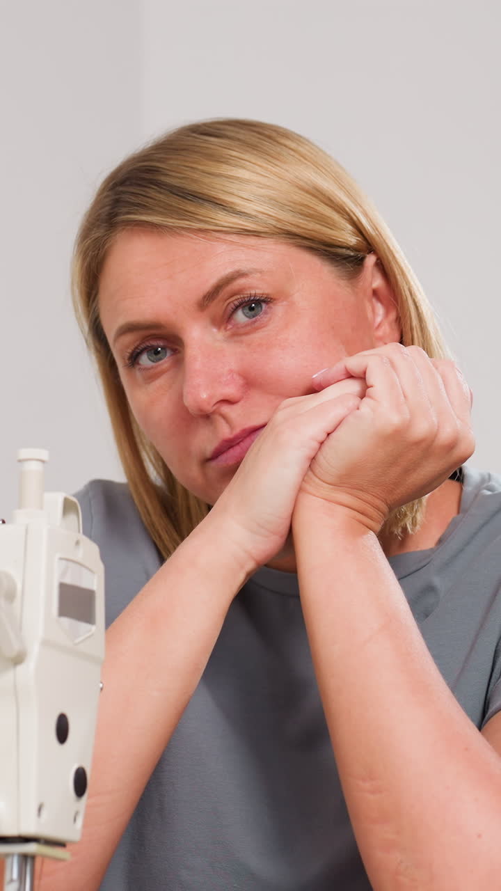 Sastre caucásico estrechando las manos cerca de la máquina, con aire contemplativo de dueño de boutique, camisa gris, iluminación de estudio, muestras de telas y notas de patrones, planificando la estrategia de pedidos con silenciosa resolución