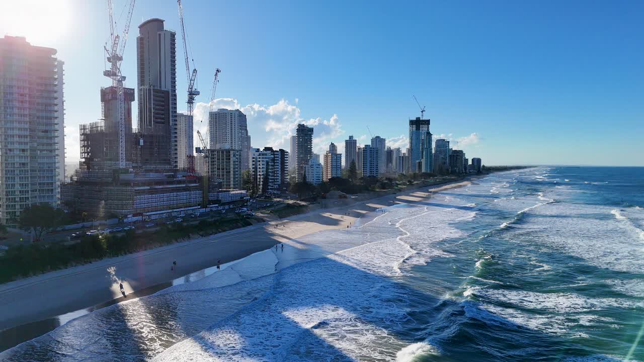 Drone footage captures the Gold Coast's vibrant skyline and ocean waves under clear blue skies, highlighting urban and natural beauty