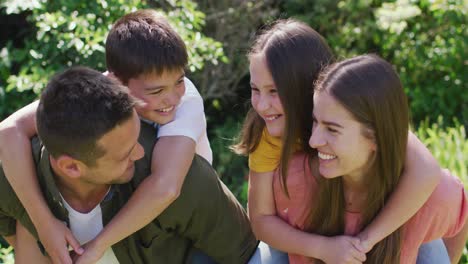 Portrait-of-caucasian-father-and-mother-carrying-son-and-daughter-on-their-back-in-the-garden