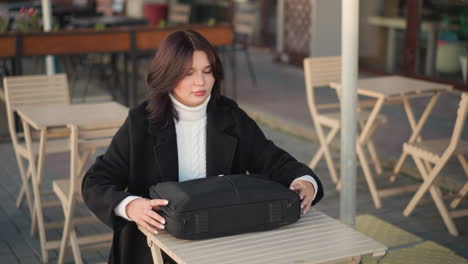 young professional seated outdoors at an urban cafe table with a laptop bag, preparing to work, stylish outfit includes black coat and white sweater, surrounded by minimalistic chairs