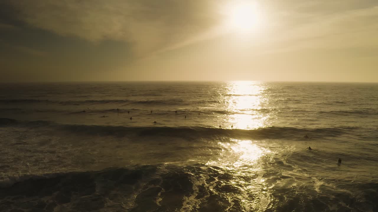 Peaceful orange sunrise aerial watching ocean waves roll onto beach with surfers silhouettes on Pacific ocean