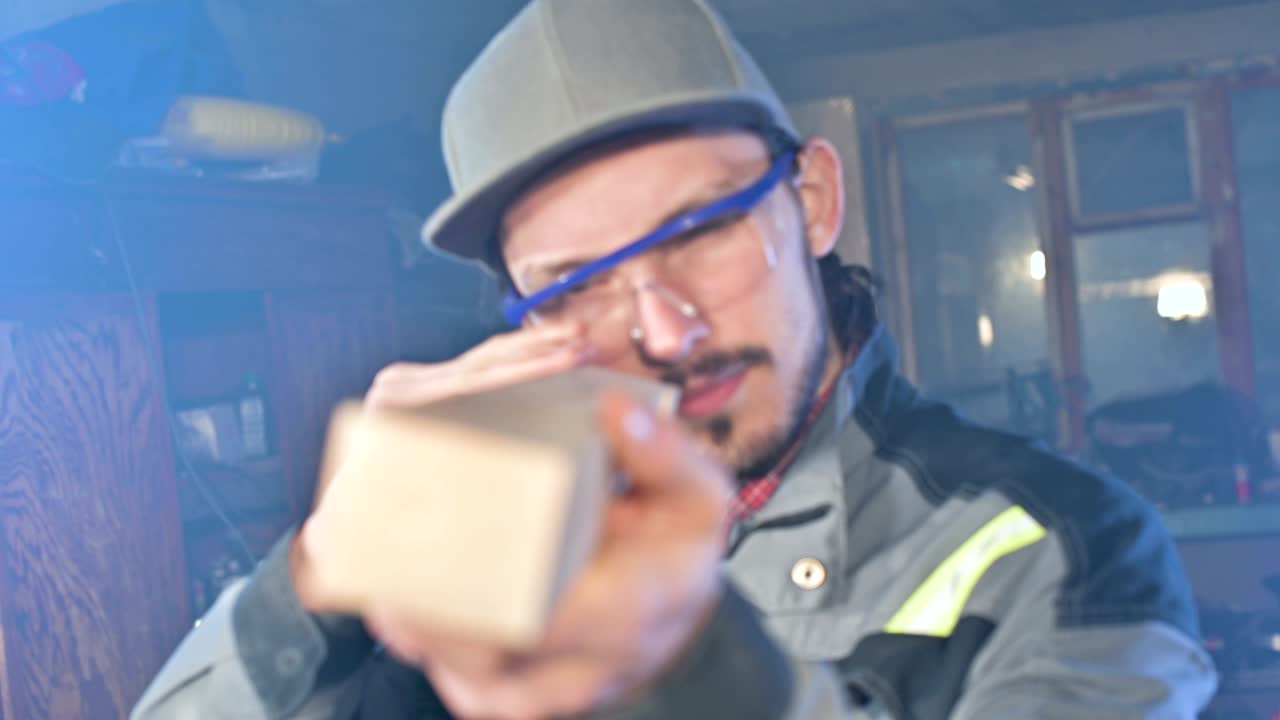 고글과 모자를 입은 집중된 젊은 수염이 있는 목공이 작업 조각을 확인합니다.