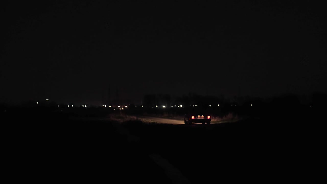 Ampia inquadratura di un'auto solitaria che scompare in lontananza su una strada di campagna vuota di notte, con i fanali posteriori che brillano nell'oscurità