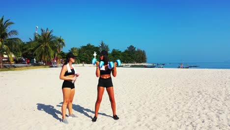 Healthy-girls-practicing-weight-lifting-on-white-sandy-beach-of-tropical-island-with-blue-sea-and-sky-background-in-Ko-Pha-Ngan,-Thailand