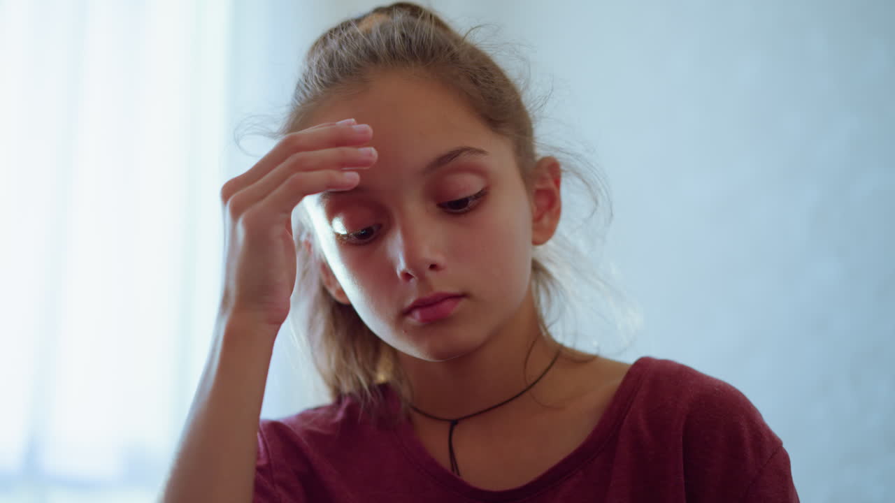 Close up of little girl with ponytail and maroon shirt resting head on hand while looking down with sober thoughtful expression in soft indoor light showing emotion