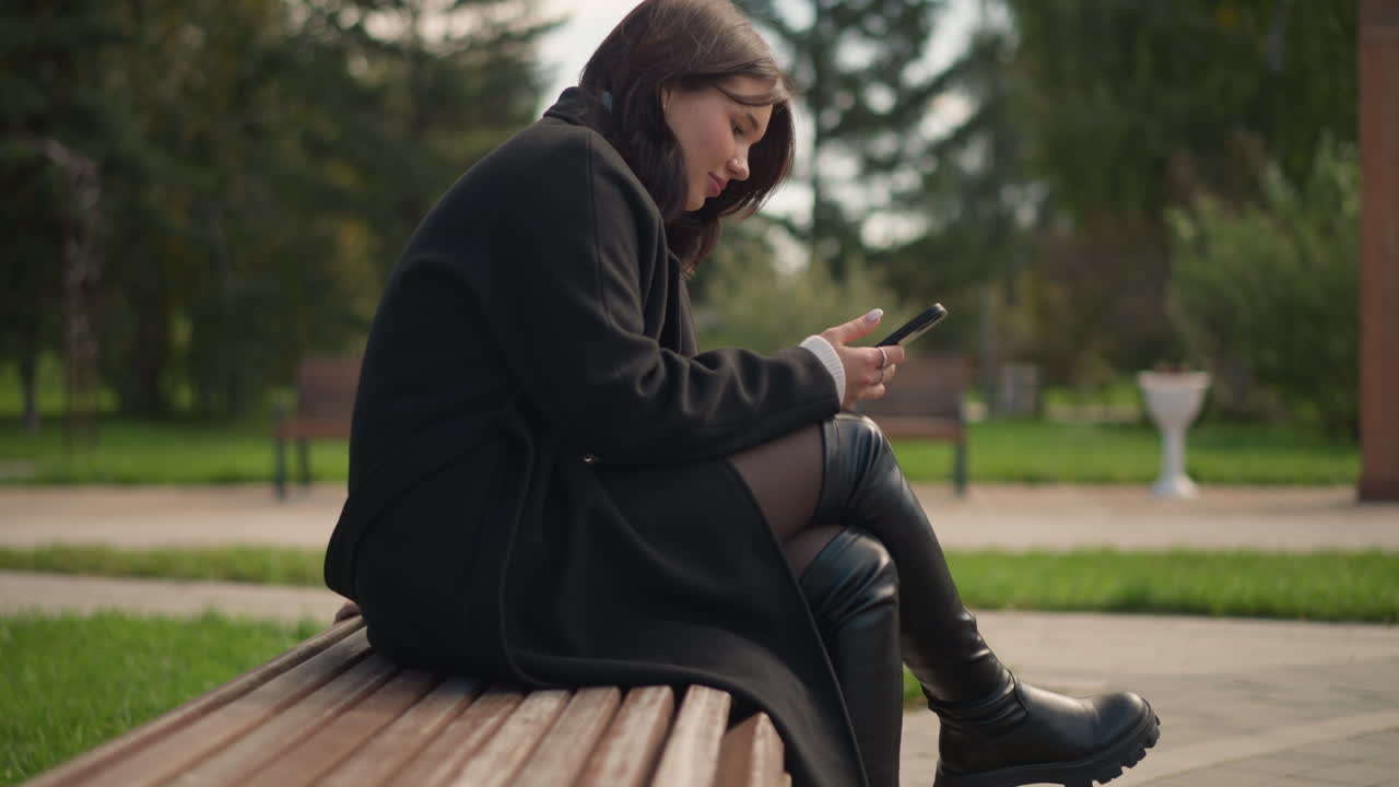 vista lateral mujer profesional en abrigo negro centrándose en su teléfono con una sonrisa sutil, sentada con las piernas cruzadas en un banco del parque, rodeada de vegetación