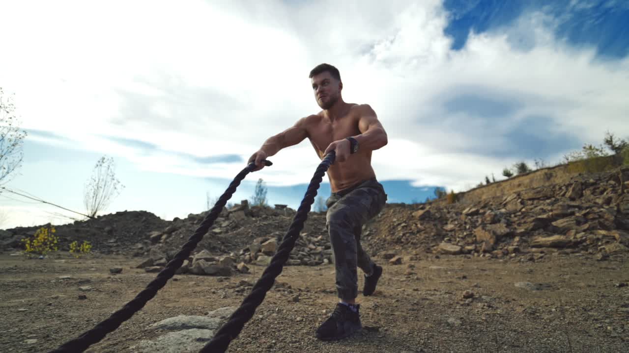 Professional sportsman working out with battle ropes. Muscular man without shirt pumping muscles with cables on the nature rocky background.