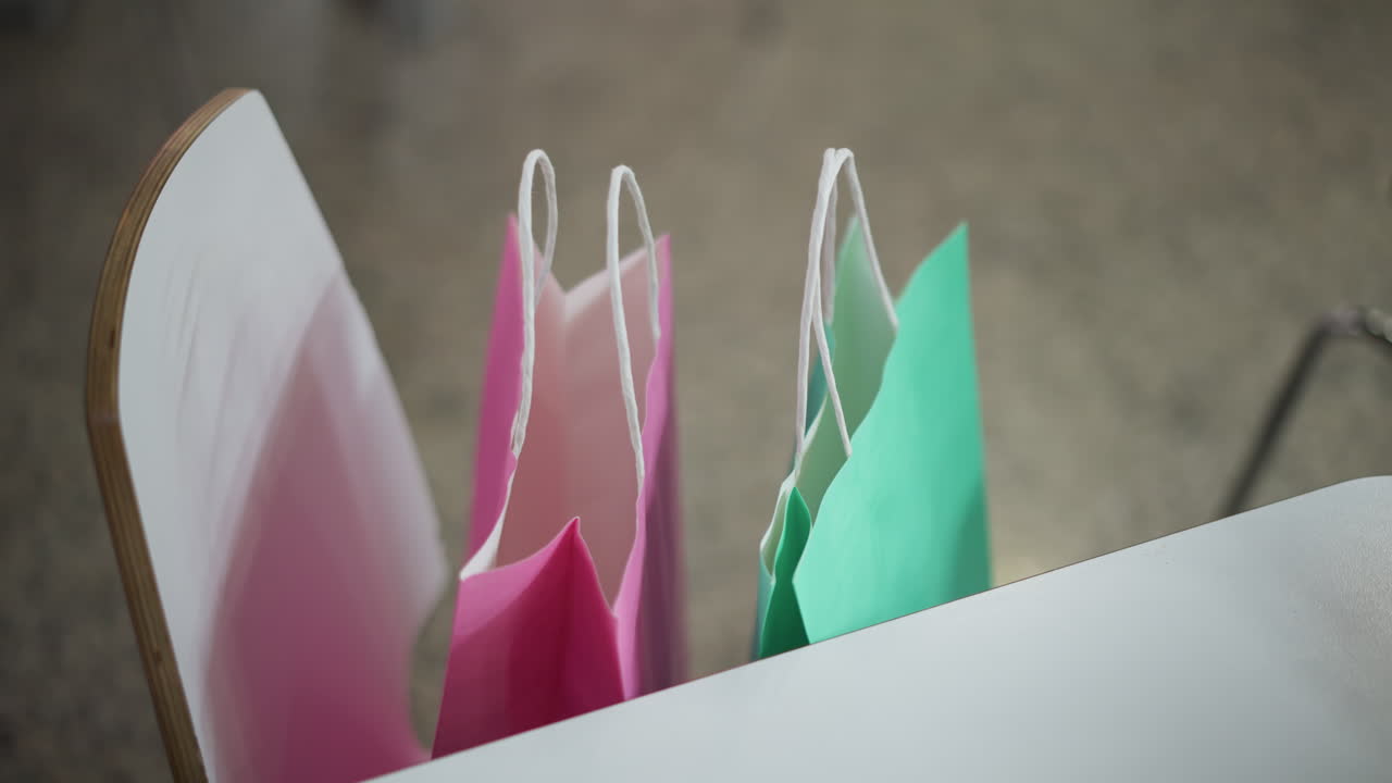 bolsas de compras rosas y verdes brillantes colocadas cuidadosamente en una silla con vista parcial de una mesa en un entorno interior moderno, mostrando un vibrante contraste de colores y simplicidad