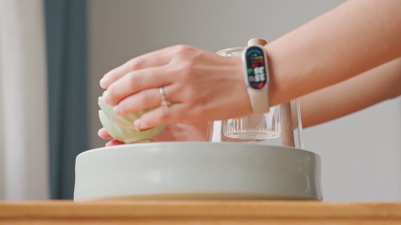 Vassoio del servizio in camera d'hotel con bicchiere e ciotola, mani posano delicatamente un bicchiere capovolto e una ciotola di ceramica su un tavolo di legno vicino alla tenda, smartwatch visibile, luce mattutina crea una scena di ospitalità minimalista.