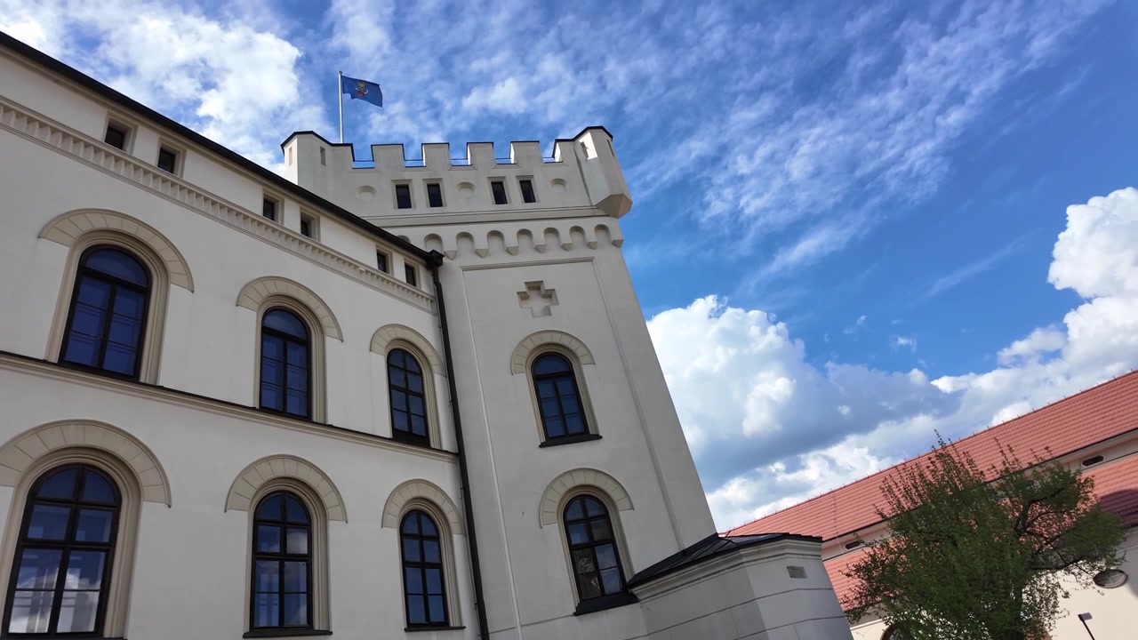 Valkoinen torni kohoaa keskiaikaisen rakennuskompleksin yläpuolelle, vasemmalla asuinrakennusten muurit ja oikealla punakattoinen talli.