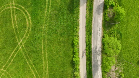 Una-Vista-Aérea-De-Un-Camino-Rural-Con-Dos-Ciclistas-Recorriéndolo,-Rodeado-De-Campos-Verdes