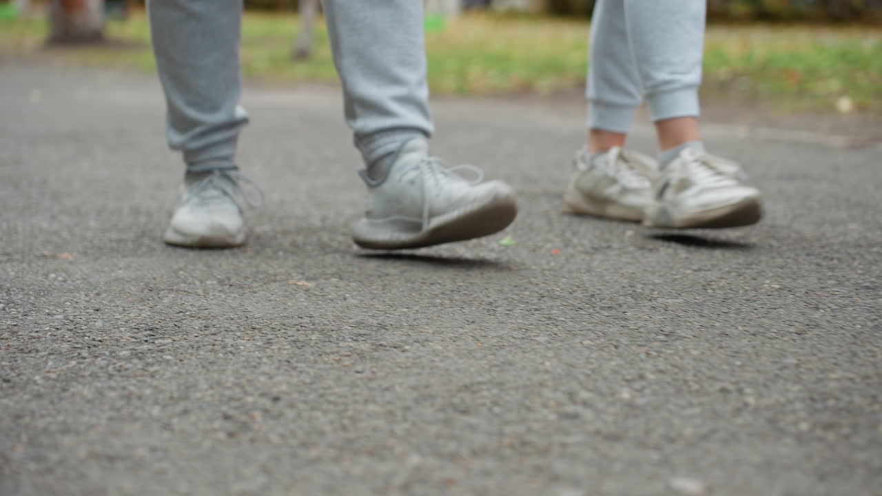 Lower angle view of two people walking side by side in grey joggers and sneakers on paved road with visible motion, relaxed stride, and outdoor setting surrounded by greenery and blurred elements