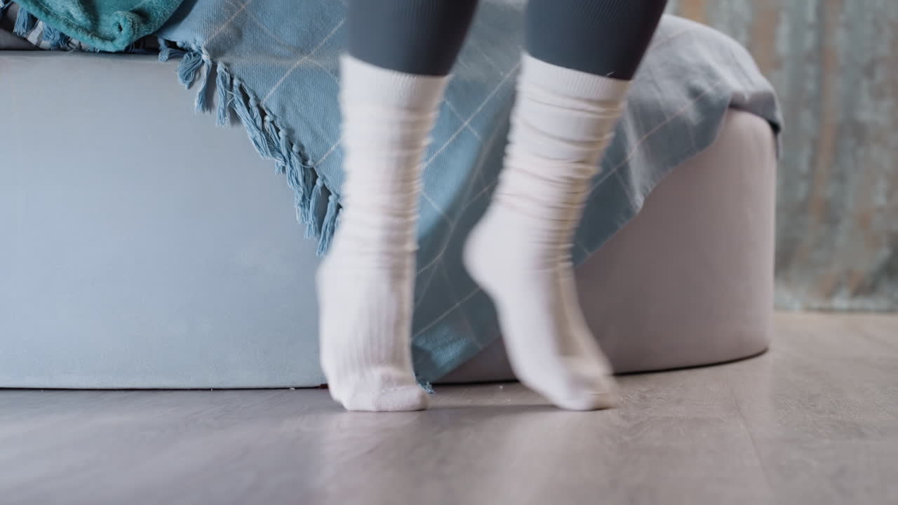 Closeup of person feet in white socks jumping on wooden floor near bed with blue blanket, cozy indoor detail showing comfort, relaxation, warmth, peaceful atmosphere
