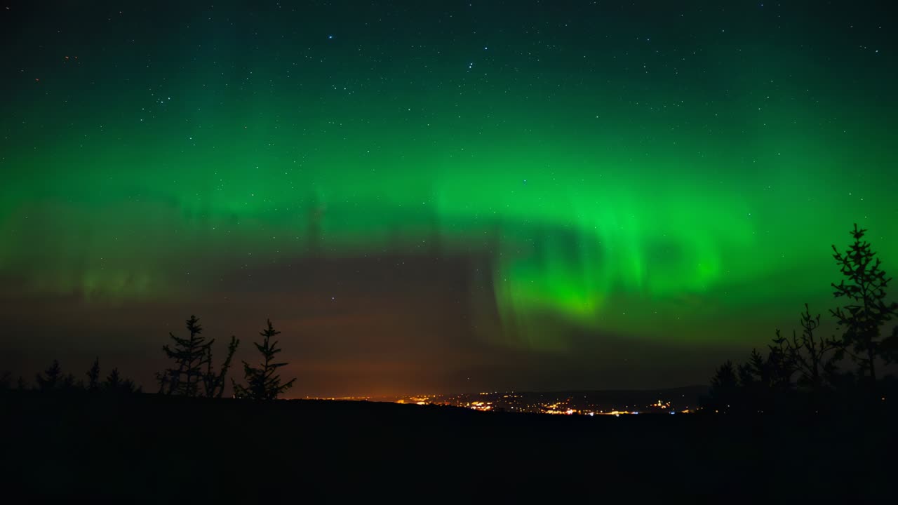 Nightfall allowing green auroral band brightening and spiraling over upland ridge, with town lights