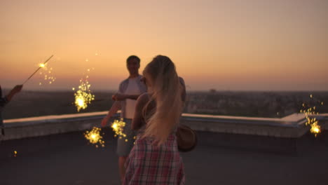 rubia rusa en el techo mueve sus brazos y cuerpo hermosamente un baile con sus amigos en una noche de verano con gran luz bengal. su cabello sopla hermosamente en el viento.
