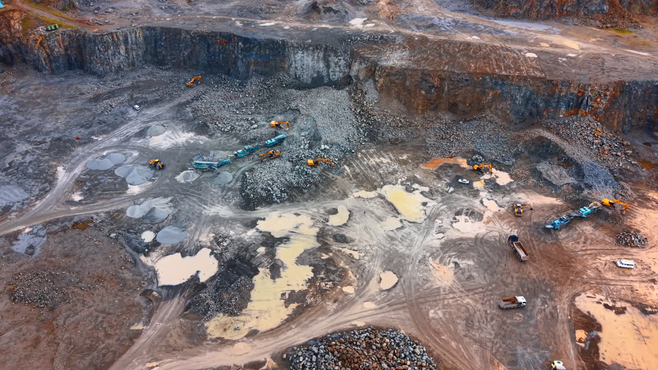 Wide aerial shot of active mining site with puddles. High angle view of a quarry floor with heavy machines, piles of stone, and rainwater puddles