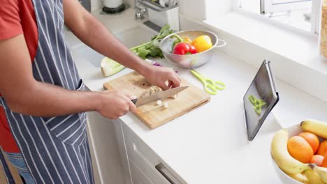 Mid-section-of-biracial-man-in-apron-chopping-vegetables,-using-tablet-in-kitchen,-slow-motion