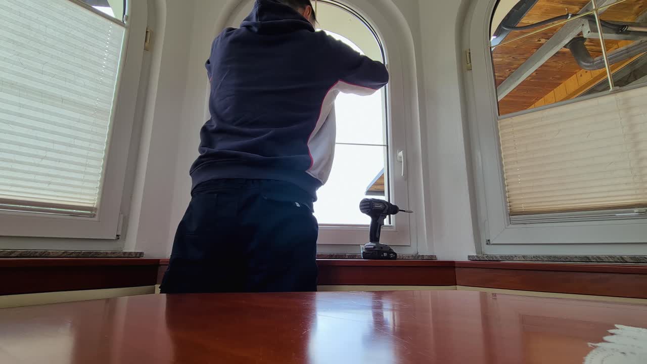 Innendørs opptak av en mann som fester plisségardiner på et vindu ved hjelp av en drill, gjør-det-selv oppussing, interiøroppsett.