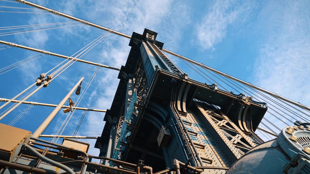 Manhattan Bridge steel tower structure, New York. Low angle perspective looking up at metal suspension framework and cables against blue sky with light clouds.