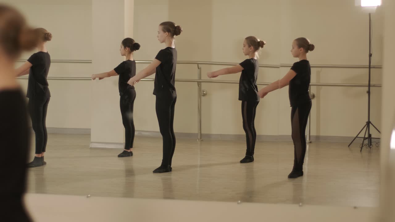 группа молодых студентов балета в черной танцевальной одежде практикует позиции в просторной балетной студии с деревянным полом и настенными решетками. сосредоточенные выражения и синхронизированные движения.