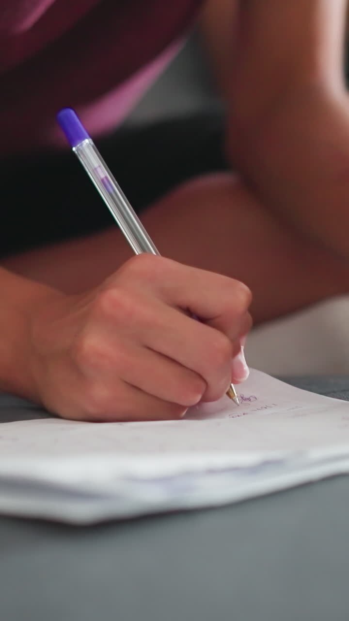 Bambina seduta a gambe incrociate sul divano che scrive su un foglio bianco con una penna blu, indossa un braccialetto di corda rosso al polso mentre la gamba di un'altra persona appare accanto a lei durante un momento di apprendimento tranquillo e concentrato