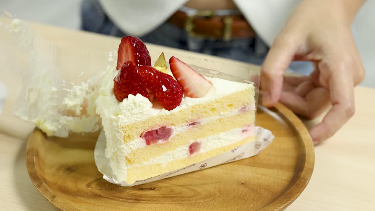 Hand carefully peels clear acetate from layered strawberry cake slice under bright, even lighting