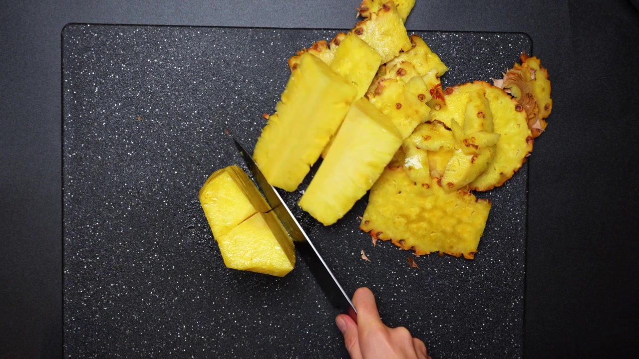 Top view of hands peeling and slicing pineapple on board. Overhead technical shot of hands using a knife to slice a fresh yellow pineapple on a black textured board.