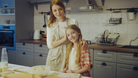 portrait shot of the young mother and her pretty daughter caressing in front of the camera and than posing in the kitchen while making a daugh. indoors