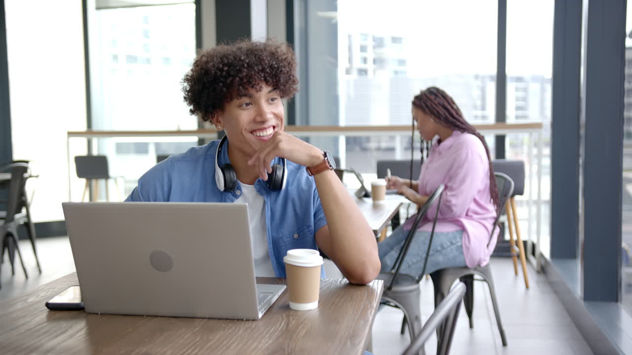 Smiling man with headphones using laptop and drinking coffee in modern cafe