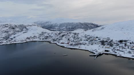 drone view in tromso area in winter flying over a fjord towards a small snowy town with small houses in norway