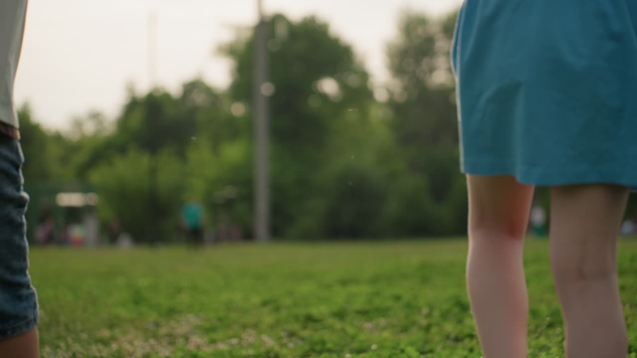 back view of boy holding hands with companion swaying while walking on green grass in park, summer light casting warm glow, casual shirt, peaceful family moment showing gentle connection and movement