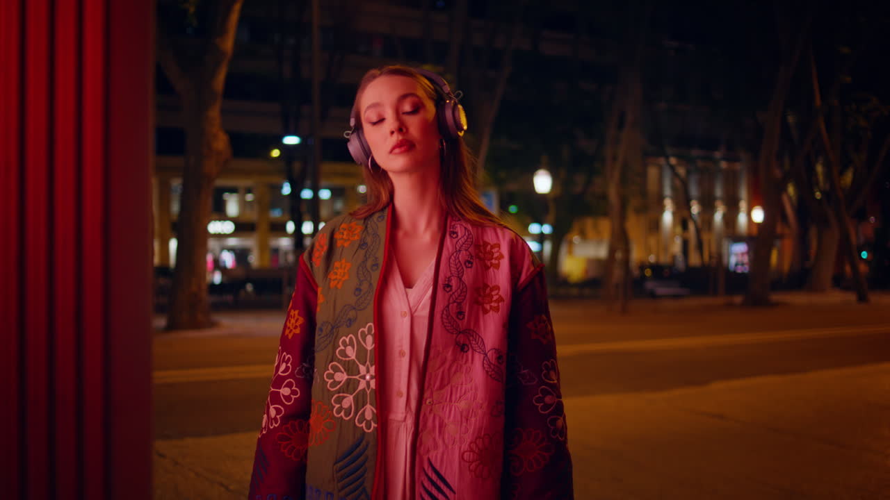 Ragazza con le cuffie in posa in una strada cittadina di notte, primo piano. Donna che ascolta musica in città.