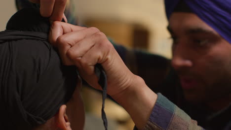 close up of father tying turban onto head of young sikh son with top knot sitting on sofa at home