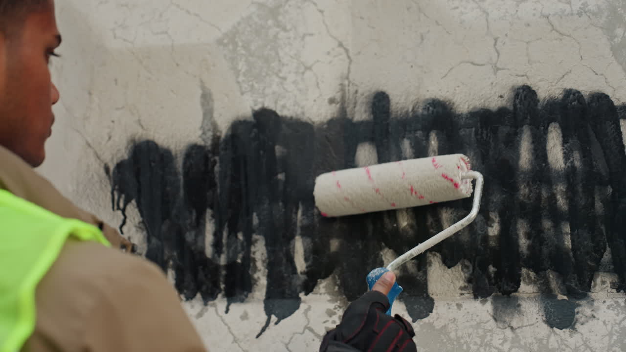 Uomo di colore che ridipinge un muro graffitato, operaio di manutenzione usa un rullo per riparare e restaurare una superficie di cemento crepata, indossando un gilet ad alta visibilità e guanti, pennellate orizzontali ferme che coprono le scritte nere.