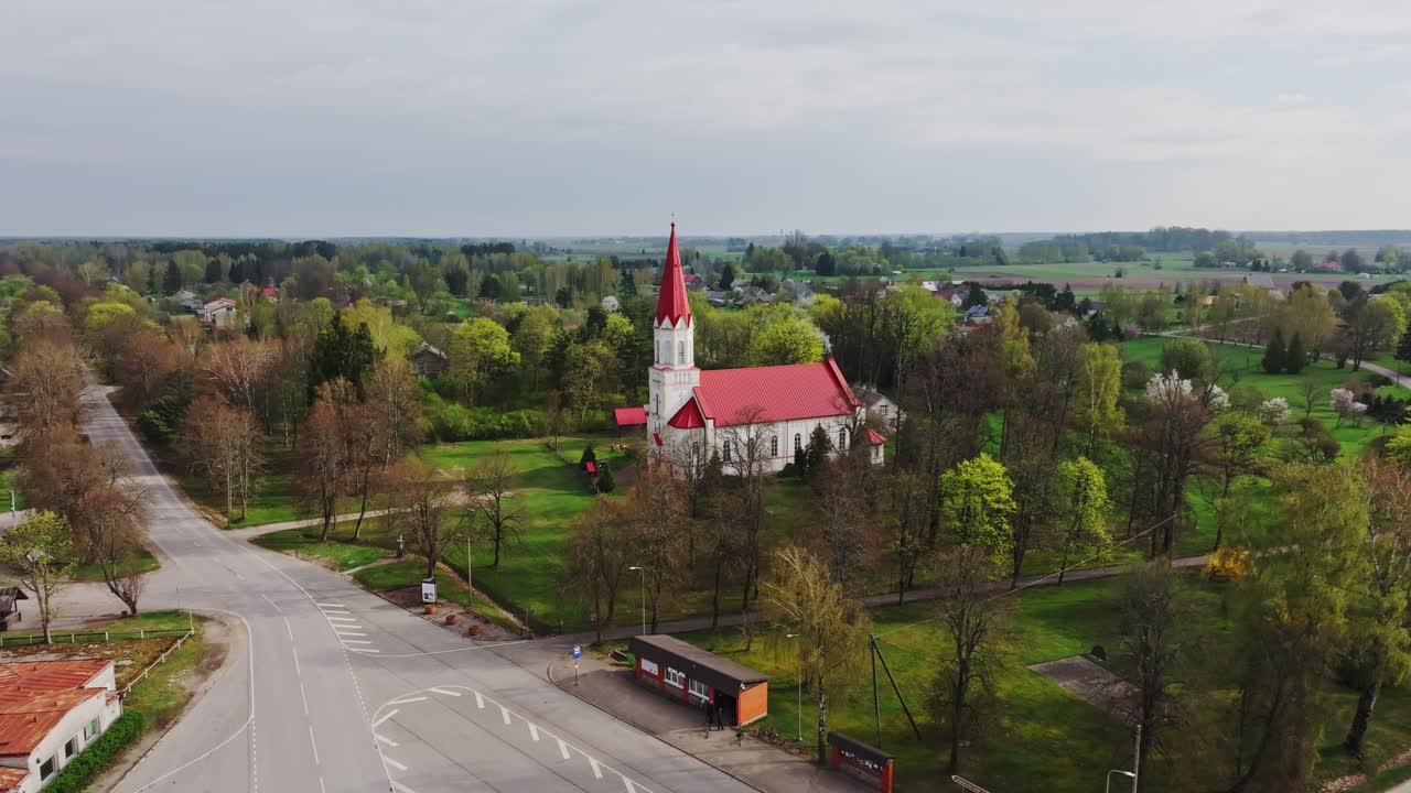 Iglesia blanca con techo rojo, revelada por el lento alejamiento de un dron, iglesia de Rucava