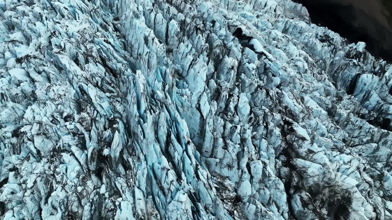 Drone flyover of a rugged Iceland glacier with blue seracs, deep crevasses, and dark volcanic ash lines across fractured frozen ridges