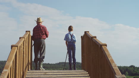 Rear-view-of-a-grandfather-fishing-with-his-little-grandson-while-standing-on-the-wooden-bridge-and-holding-rods-in-the-water